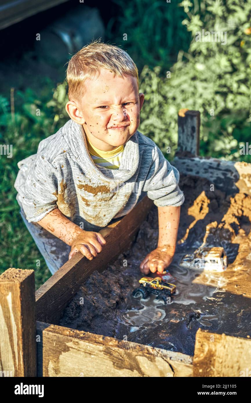 Carino ragazzo preschooler serio con il viso sporco e vestiti che giocano con le automobili giocattolo nel sandbox vicino casa rurale in legno nel giorno d'estate in campagna Foto Stock