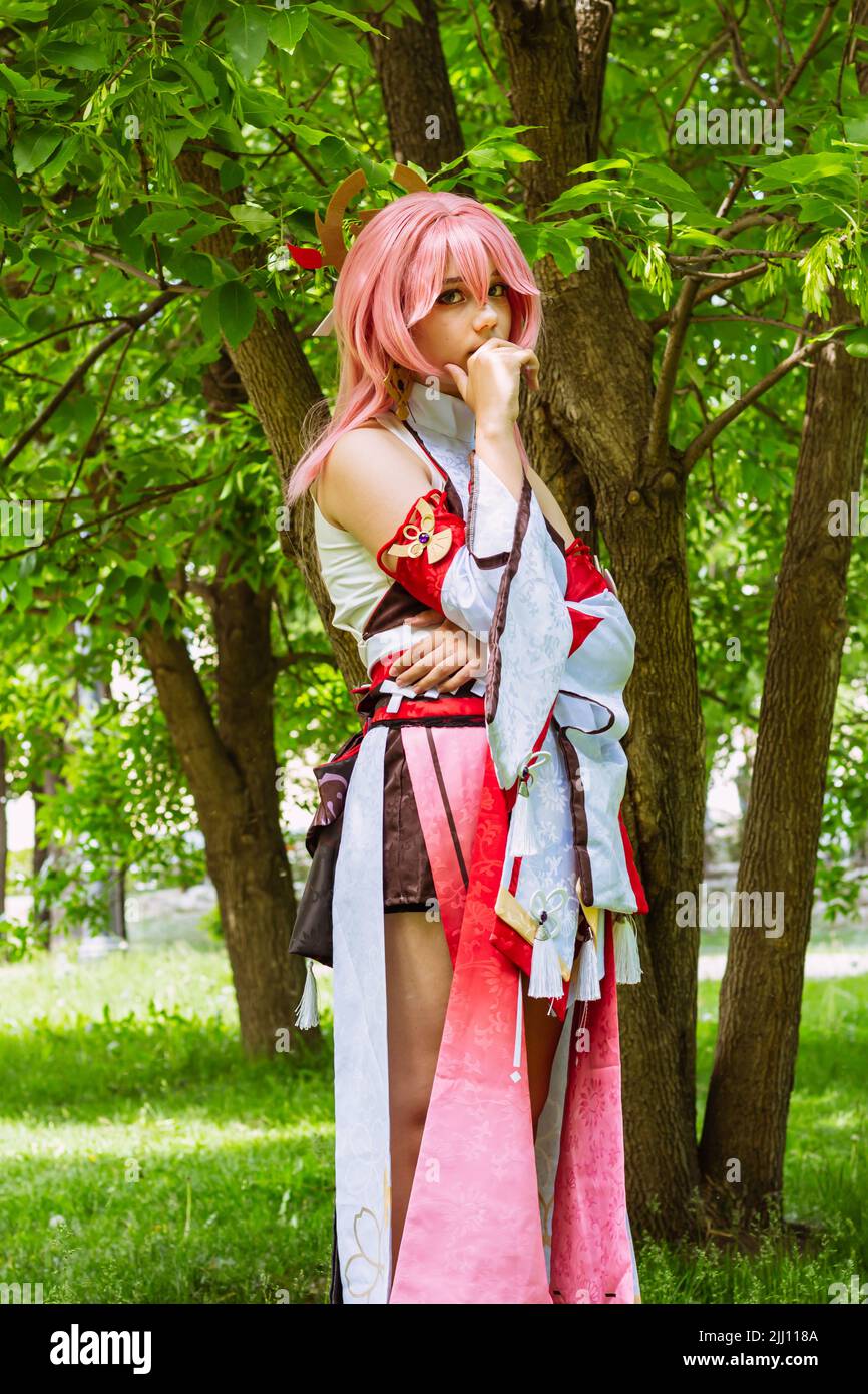 costume anime nel parco all'aperto. Personaggio della dea del gioco del computer con capelli rosa YaeMiko. Ragazza vestita come eroe anime si erge vicino alla holida estate albero Foto Stock