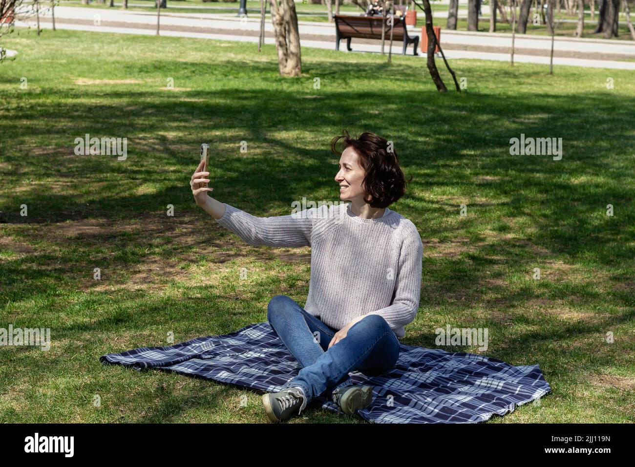 Donna sorridente attraente che fa selfie foto su smartphone su sfondo naturale, all'aperto nel parco della città seduta su plaid, fuoco selettivo Foto Stock