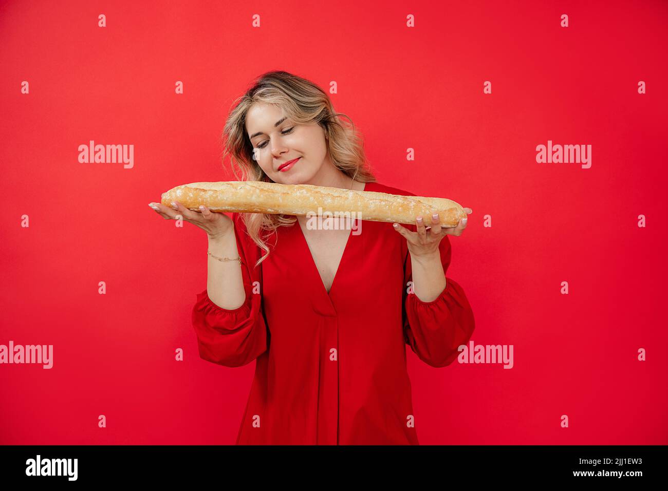 Sorridente calma attraente donna bionda in vestiti rossi tenere delicatamente e odore di pane francese baguette nelle mani in studio rosso Foto Stock