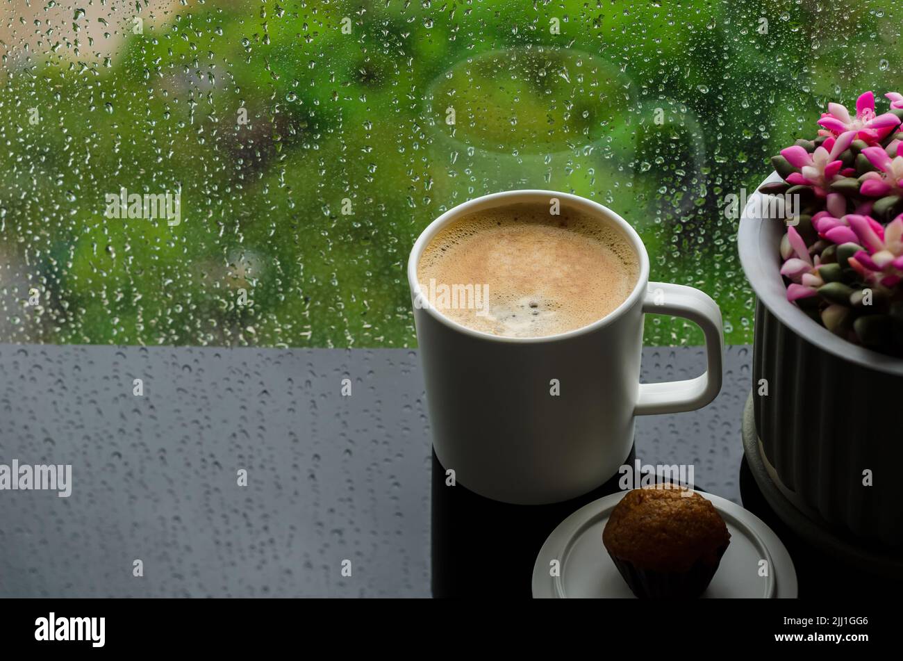 Una tazza di caffè caldo con una piccola torta al tavolo al mattino con gocce di pioggia sulla finestra e sfondo verde. Soggiorno a casa e concetto di relax. Foto Stock