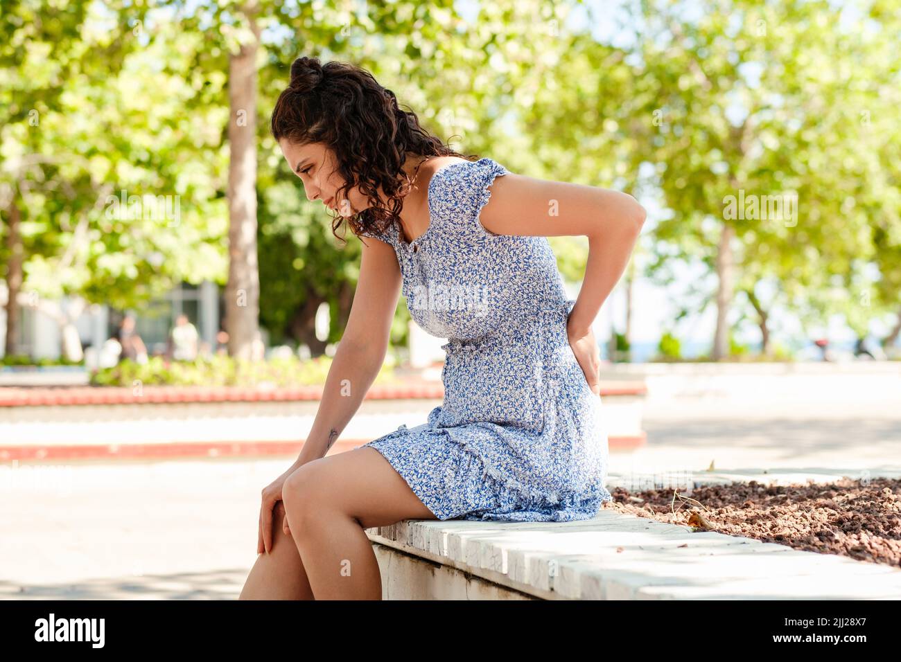 Giovane donna bruna vestito estivo sul parco cittadino, all'aperto mal di schiena soffrono di dolore lombare inferiore dolore muscolare. Massaggiare i muscoli tesi. Foto Stock