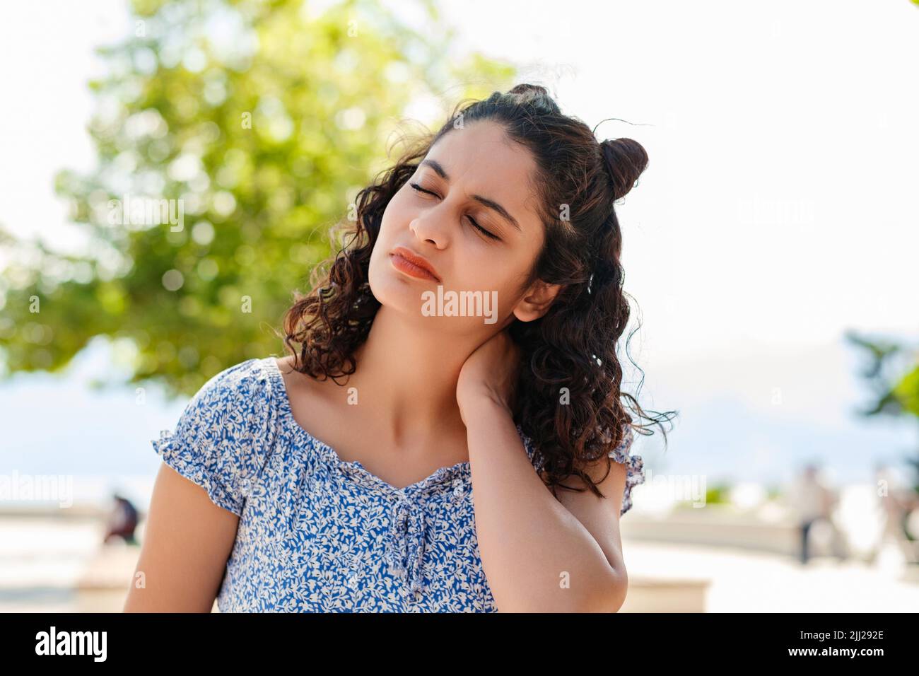 Ritratto di giovane donna bruna vestito estivo in piedi sul parco della città, all'aperto sensazione di dolore alle spalle articolare dolore alla schiena, concetto di fibromialgia Foto Stock