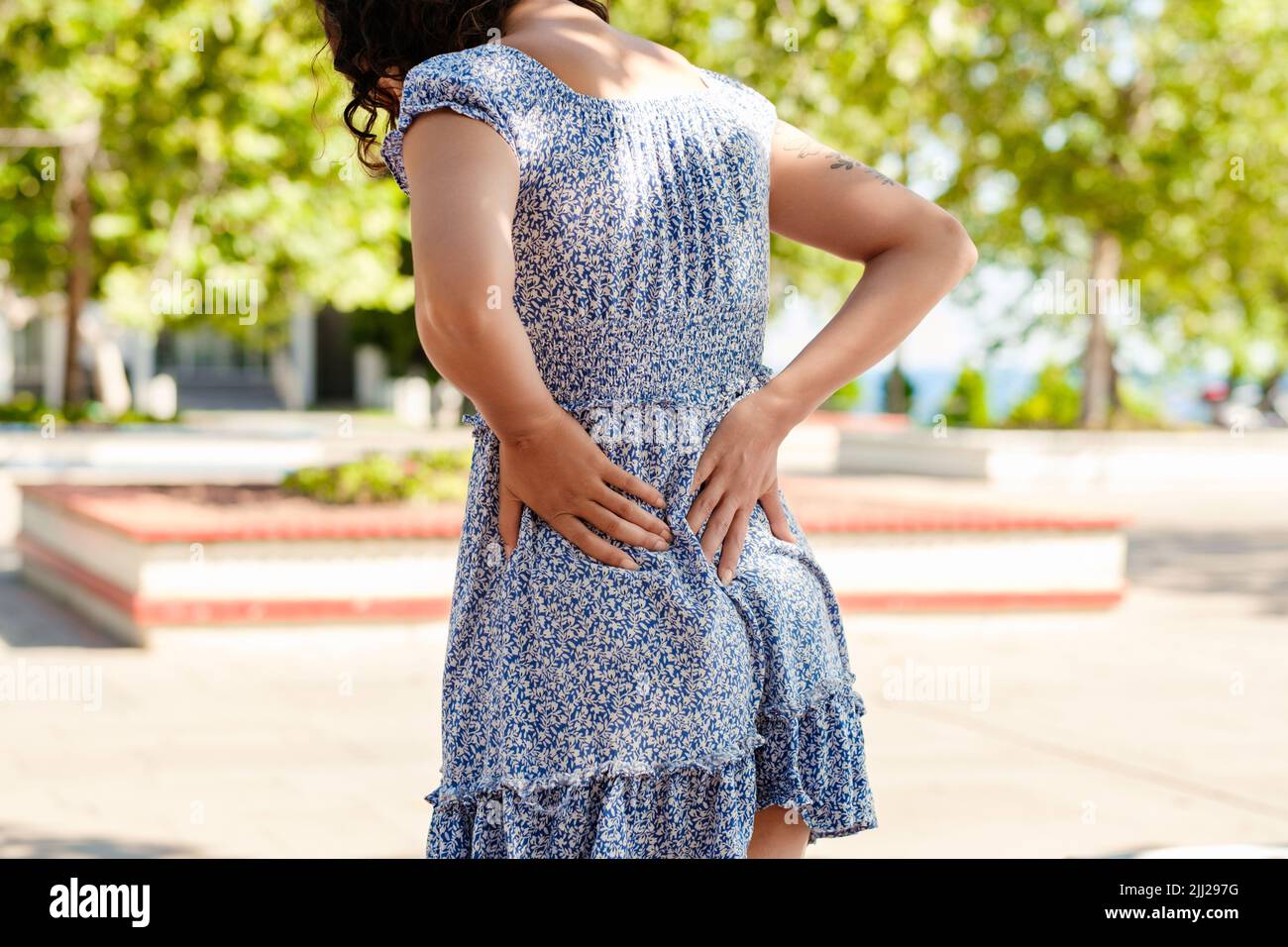 Giovane donna caucasica che indossa un abito estivo floreale blu in piedi sul parco della città, all'aperto mal di schiena soffrono di dolore lombare inferiore. Ma Foto Stock