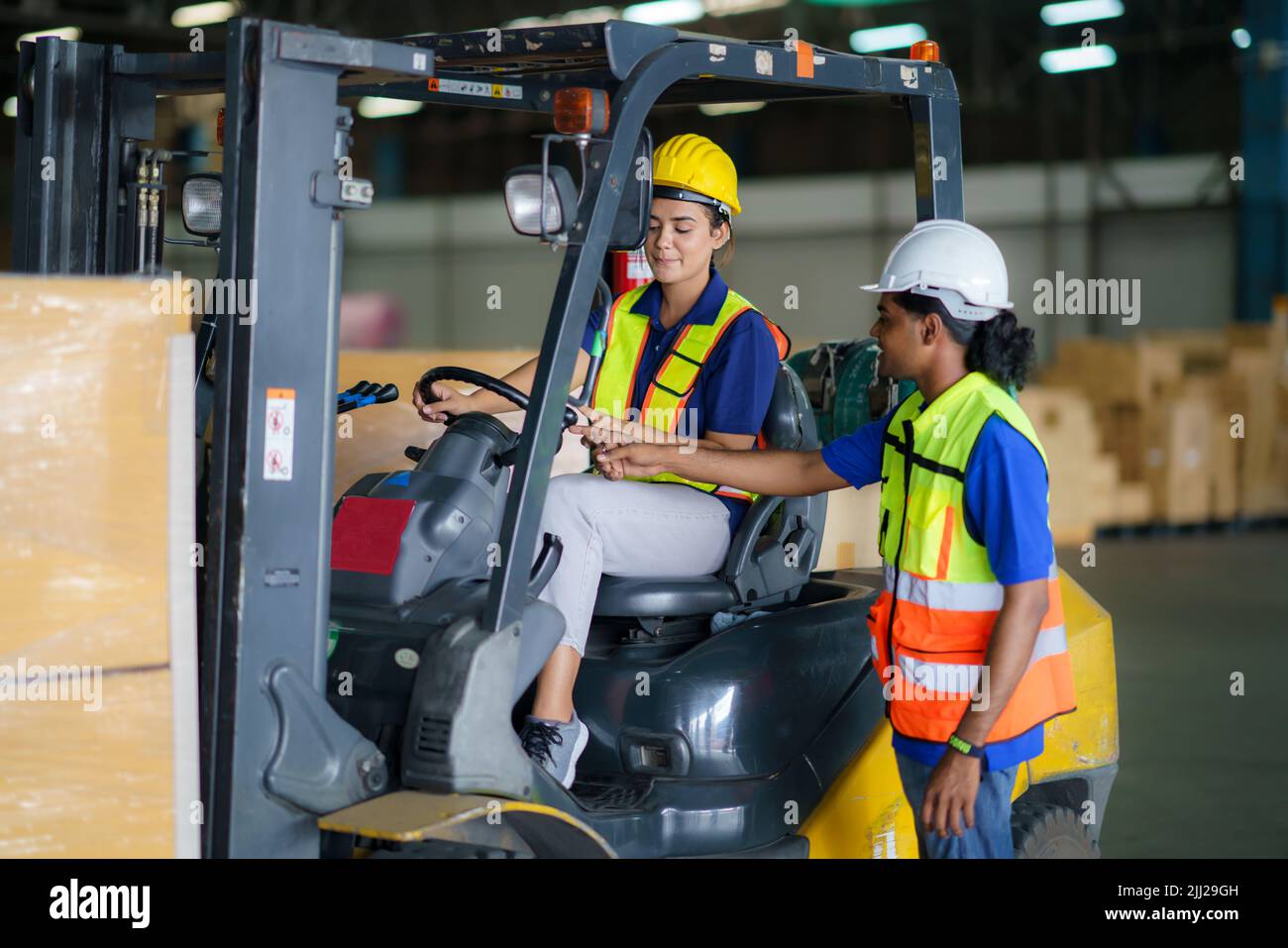 Uomo asiatico insegnamento donna conducente guida carrello elevatore auto in industria con sorriso, capacità di ragazza e diversità di carriera indossare casco in giubbotto riflettente Foto Stock