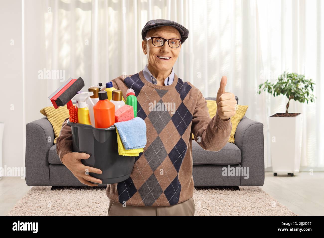 Uomo anziano con un secchio di forniture di pulizia in piedi in un soggiorno e gesturando i pollici in su Foto Stock