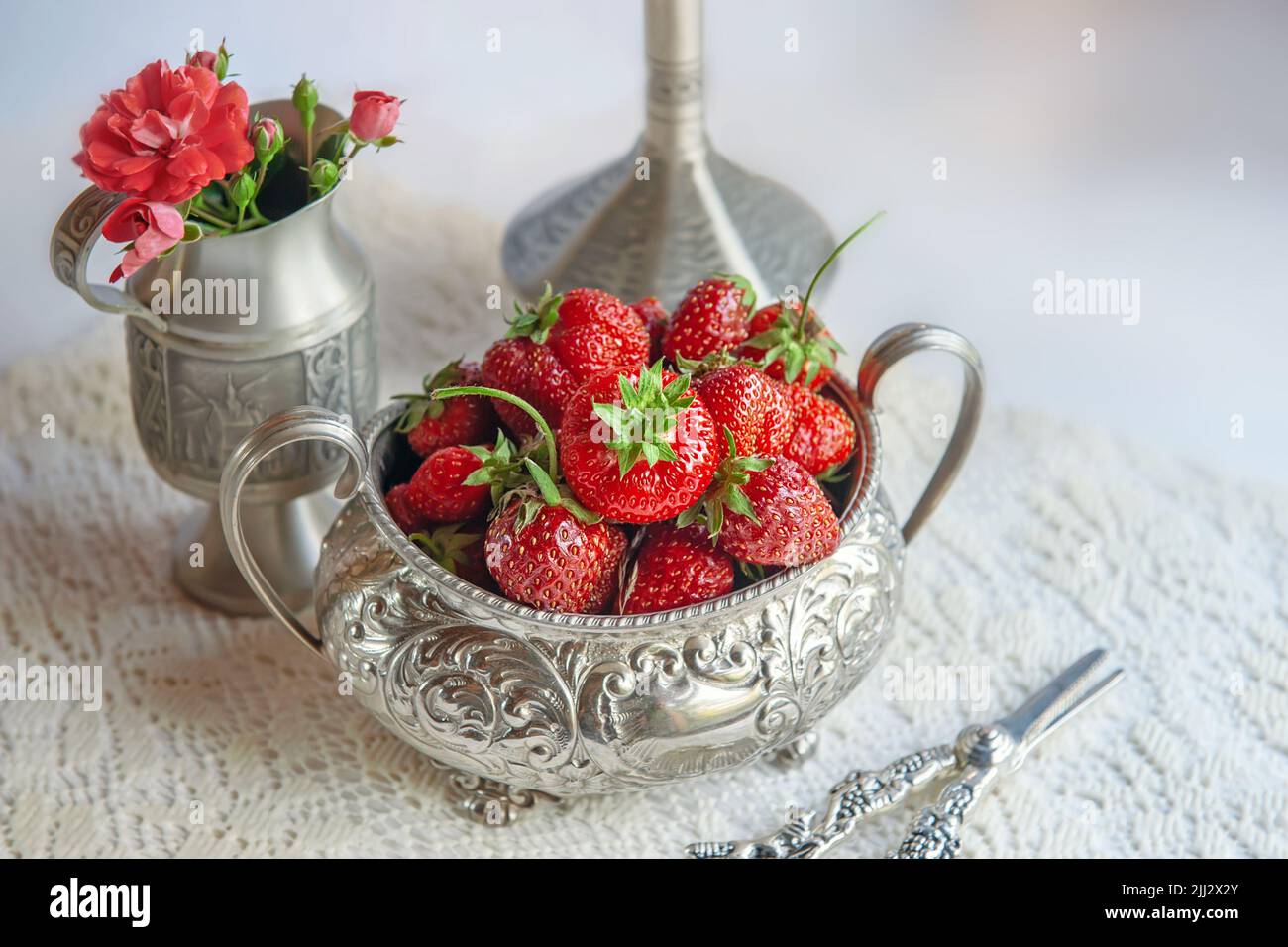 Fragole in vaso d'argento sul tavolo in stile romantico. Ancora vita di fragole, un bouquet di rose e una brocca. Foto Stock
