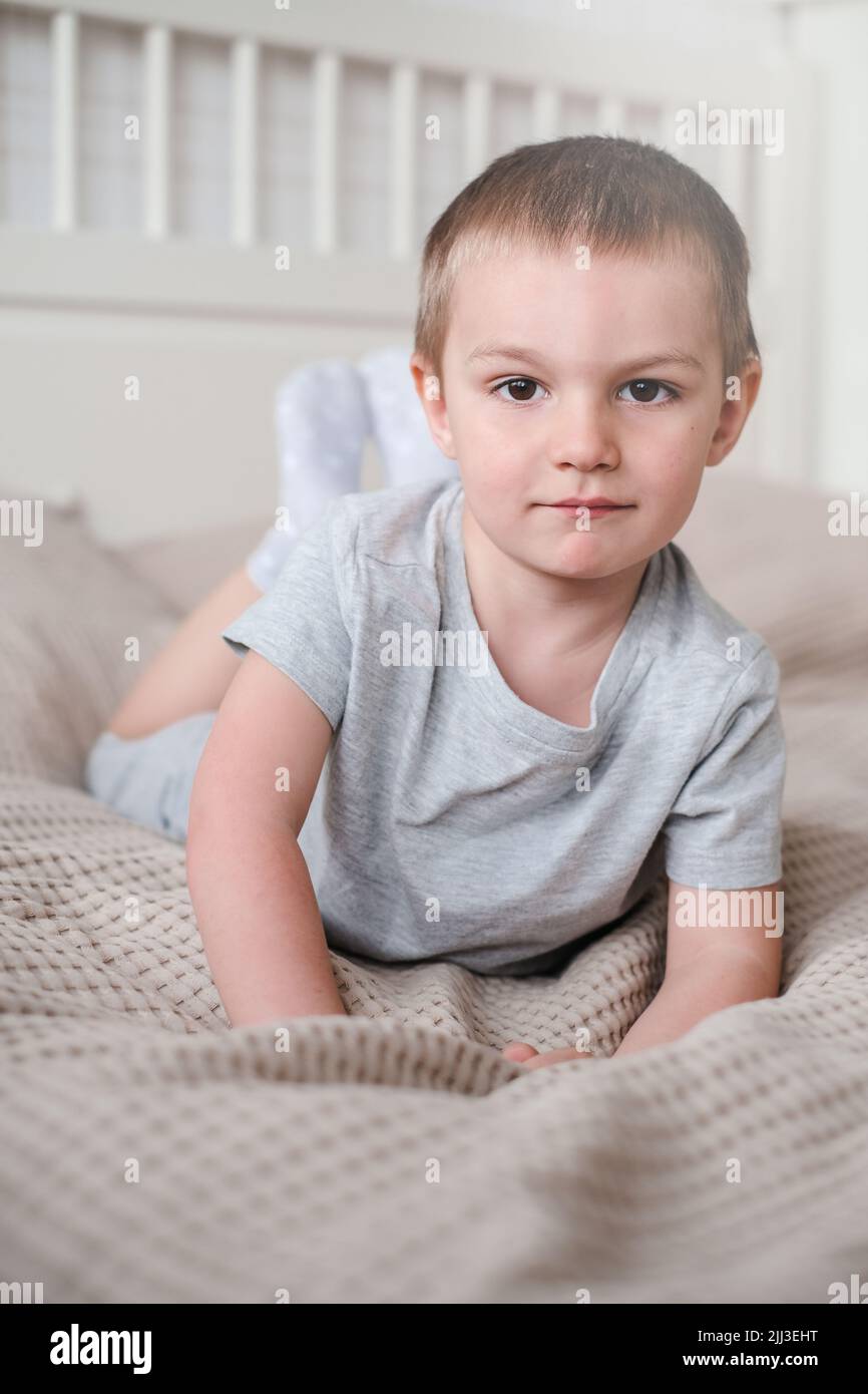 Primo piano di un piccolo ragazzo caucasico felice in una t-shirt grigia si trova sul letto e sorride Foto Stock