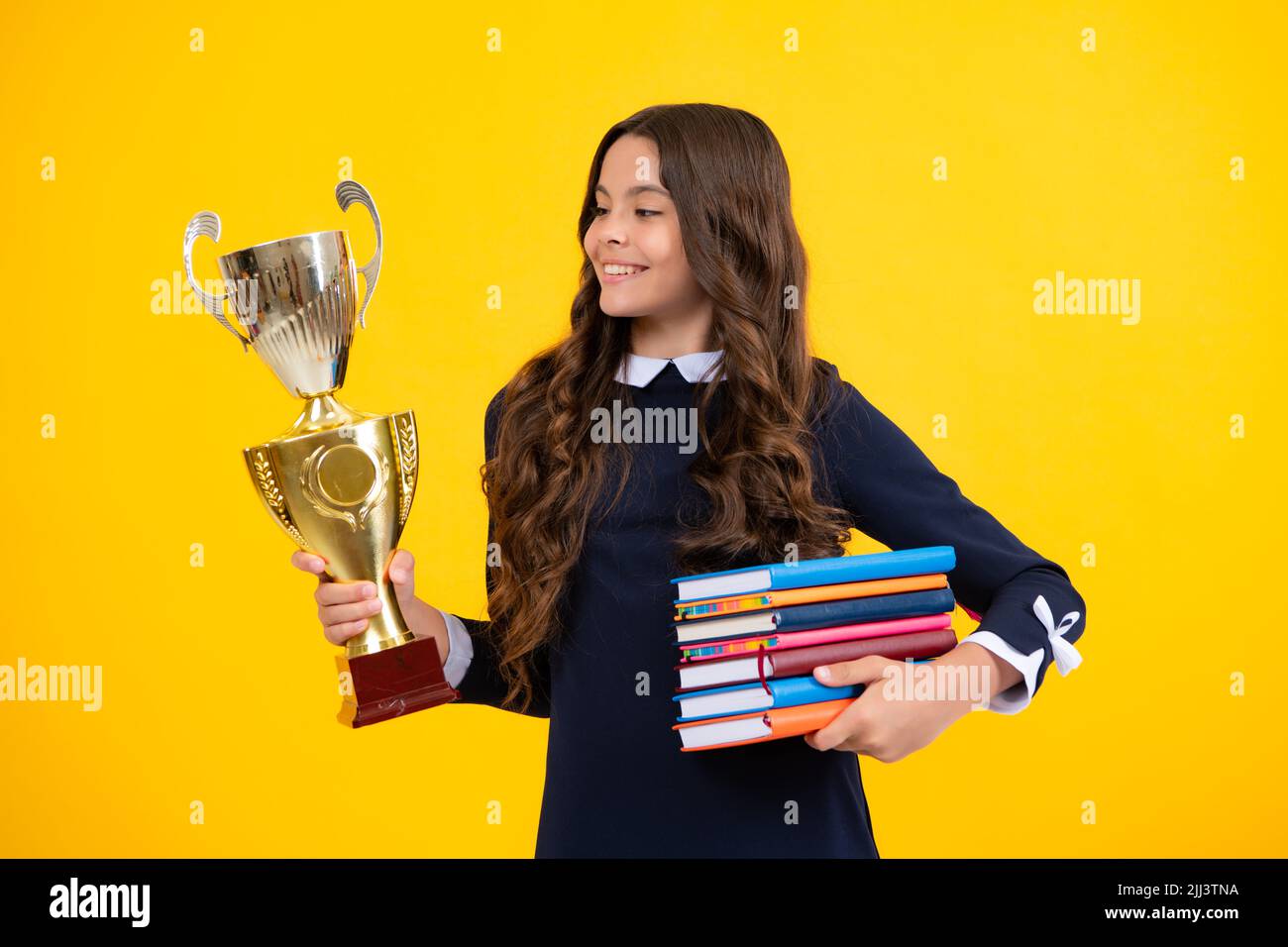 Studentessa in divisa scolastica che celebra la vittoria con il trofeo. Premio vincente per l'adolescente su sfondo giallo. Libri per bambini con trofeo d'oro Foto Stock