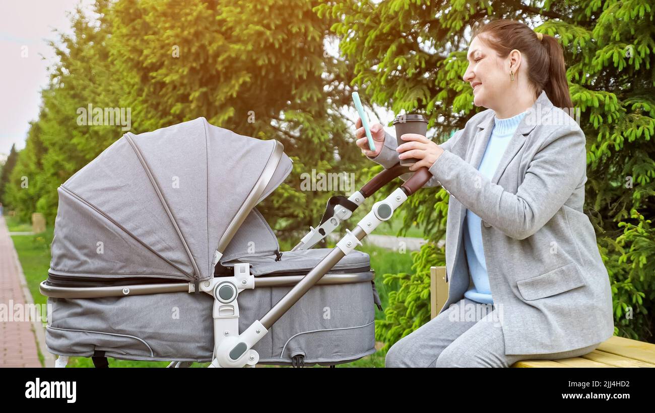 Donna rocce bambino in passeggino tenere telefono e tazza di caffè Foto Stock