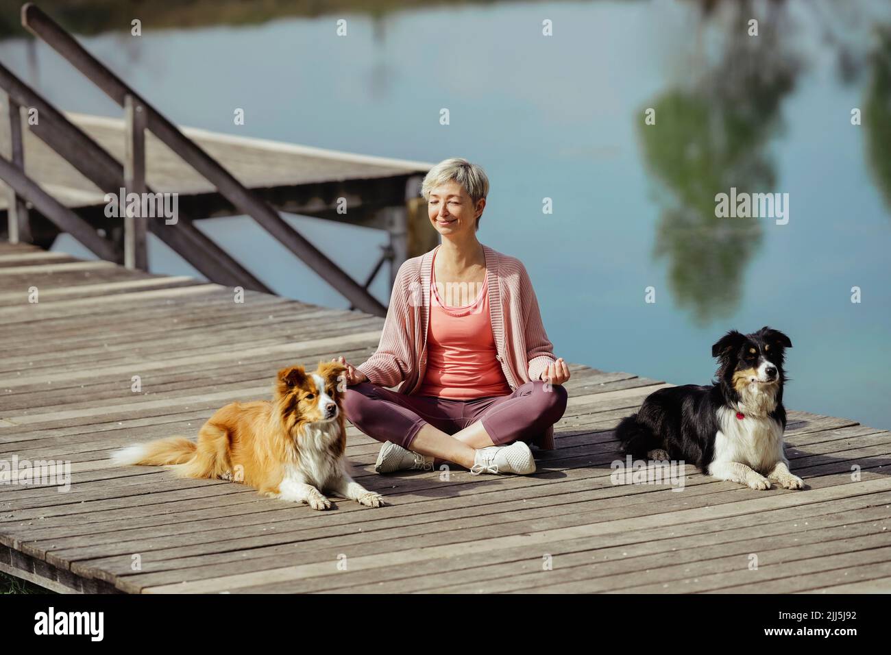 Donna matura sorridente con occhi chiusi meditando tra cani sul molo Foto Stock