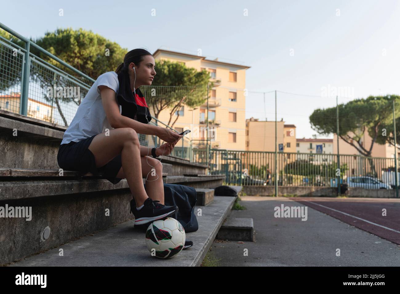 Donna con pallone da calcio che ascolta musica tramite cuffie intrauricolari sul banco del campo sportivo Foto Stock