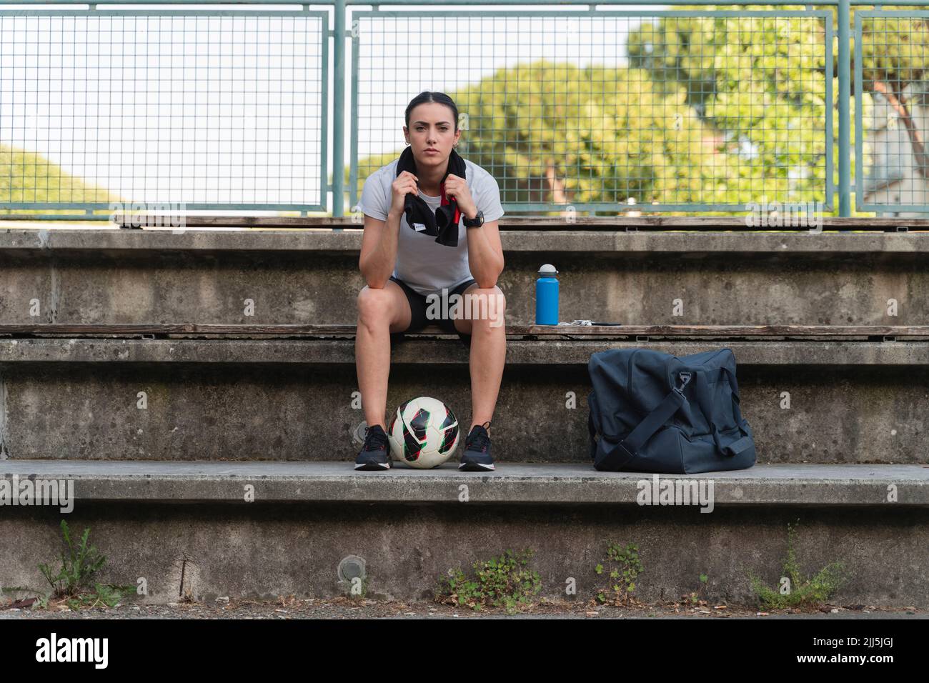 Giovane donna fiduciosa con pallone da calcio seduto su panca presso il campo sportivo Foto Stock
