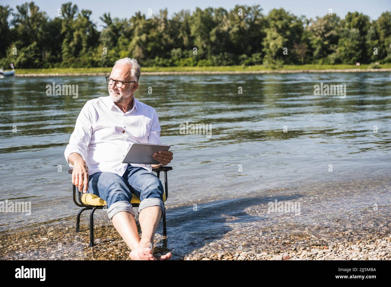 Uomo anziano che tiene un tablet PC seduto su una sedia sul lungofiume Foto Stock