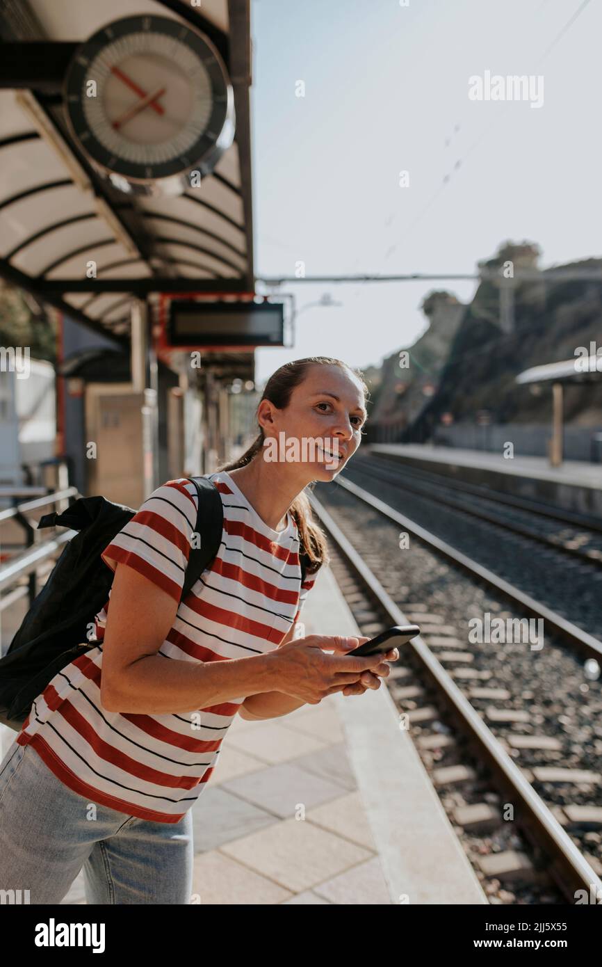 Donna sorridente con telefono in attesa alla piattaforma ferroviaria Foto Stock