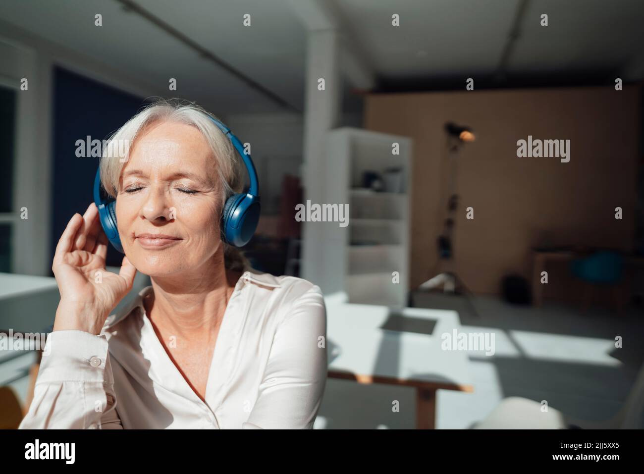 Sorridente donna anziana con occhi chiusi ad ascoltare musica attraverso cuffie wireless in ufficio Foto Stock