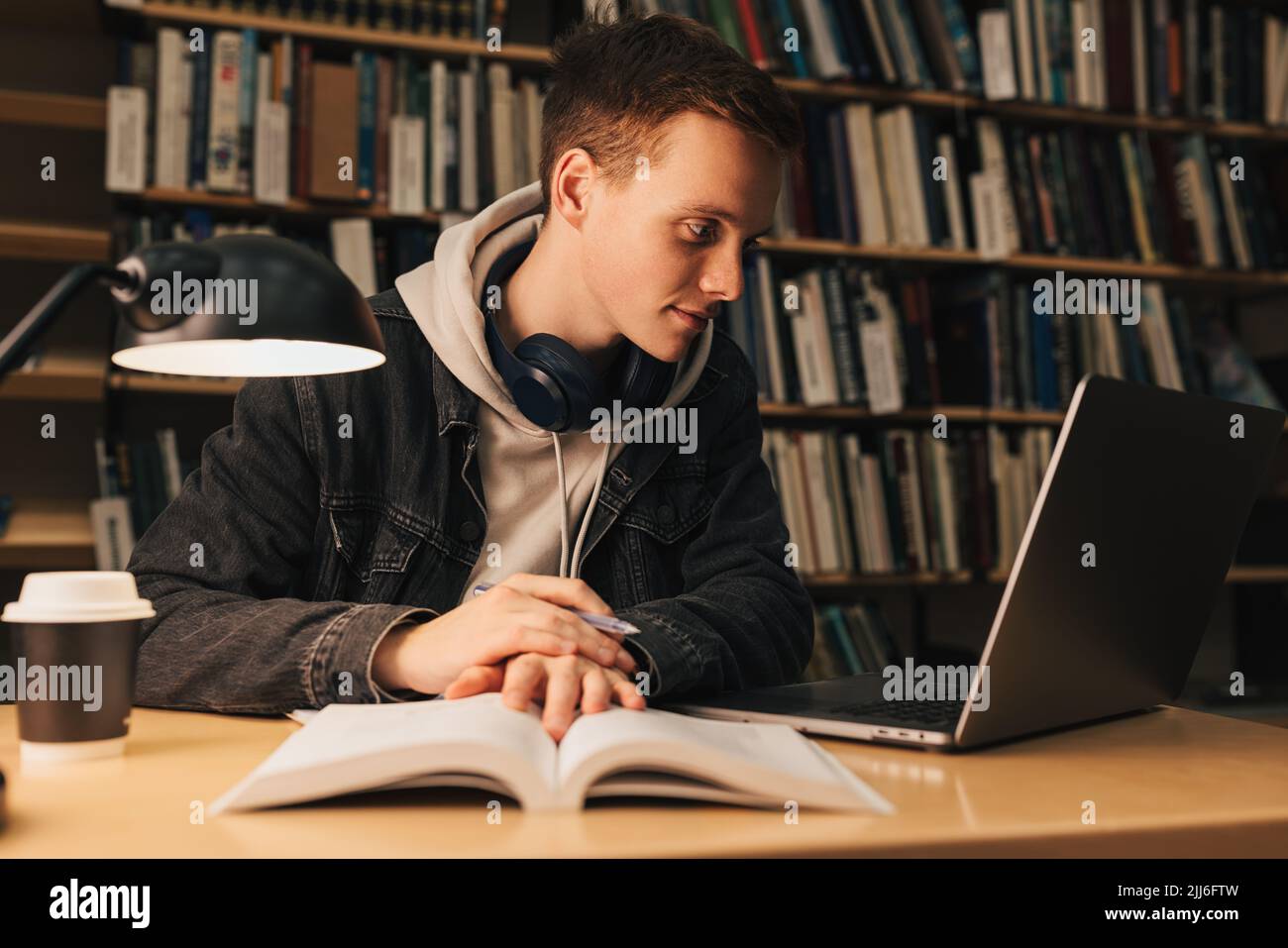 Studente maschile in biblioteca guardando il laptop. Giovane seduto alla scrivania che prepara gli esami la sera. Foto Stock