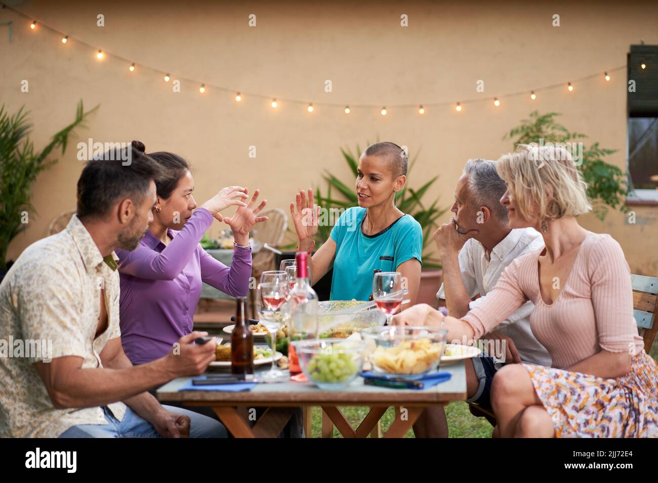 Gruppo di amici che chiacchierano durante il pranzo nel cortile. Persone di mezza età che hanno una discussione all'aperto a tavola mentre mangiano e bevono Foto Stock