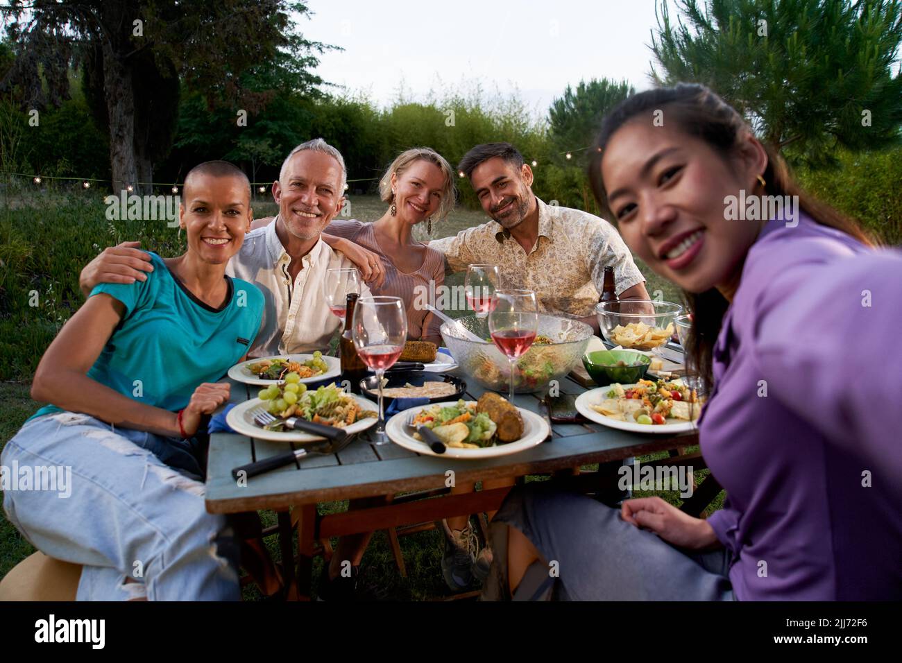 Gruppo di amici che si divertono durante la festa estiva. Donna asiatica che prende selfie all'ora della cena del barbecue. Persone di mezza età che gelano fuori mangiare e. Foto Stock