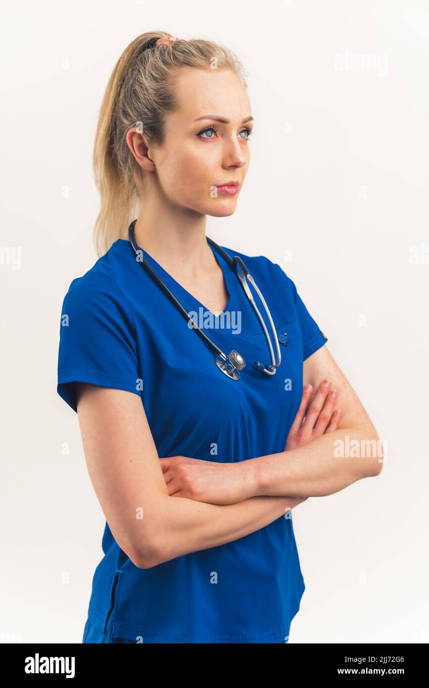 Colpo verticale di una donna sicura e forte medico. Bella donna caucasica con capelli biondi legati in uniforme clinica blu scuro e stetoscopio. Braccia incrociate, espressione facciale positiva. Sfondo bianco, spazio di copia isolato. Foto di alta qualità Foto Stock