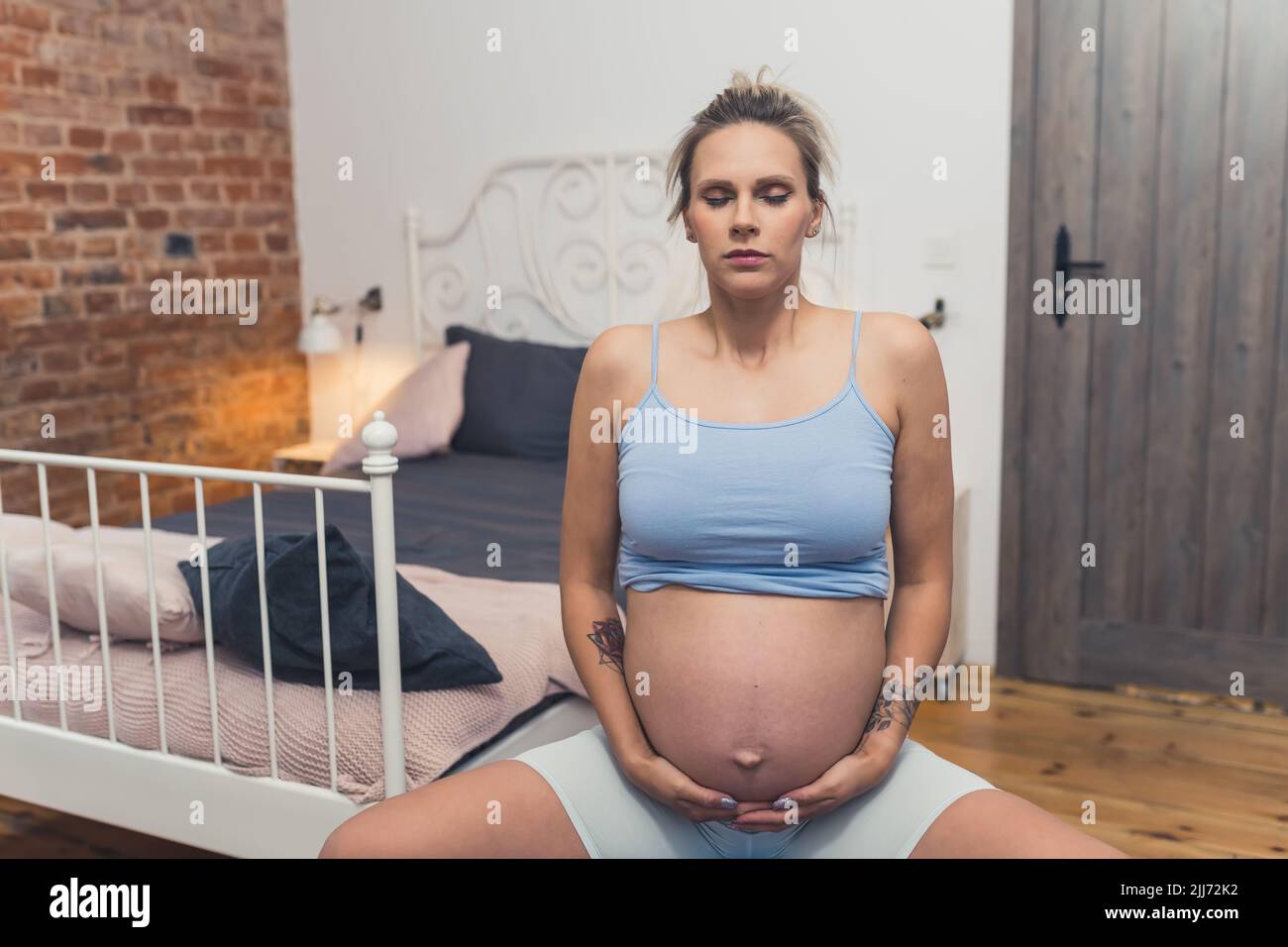 Giovane madre caucasica incinta che tocca la sua pancia e si esercita su una palla fitness. Camera da letto interna. Foto di alta qualità Foto Stock
