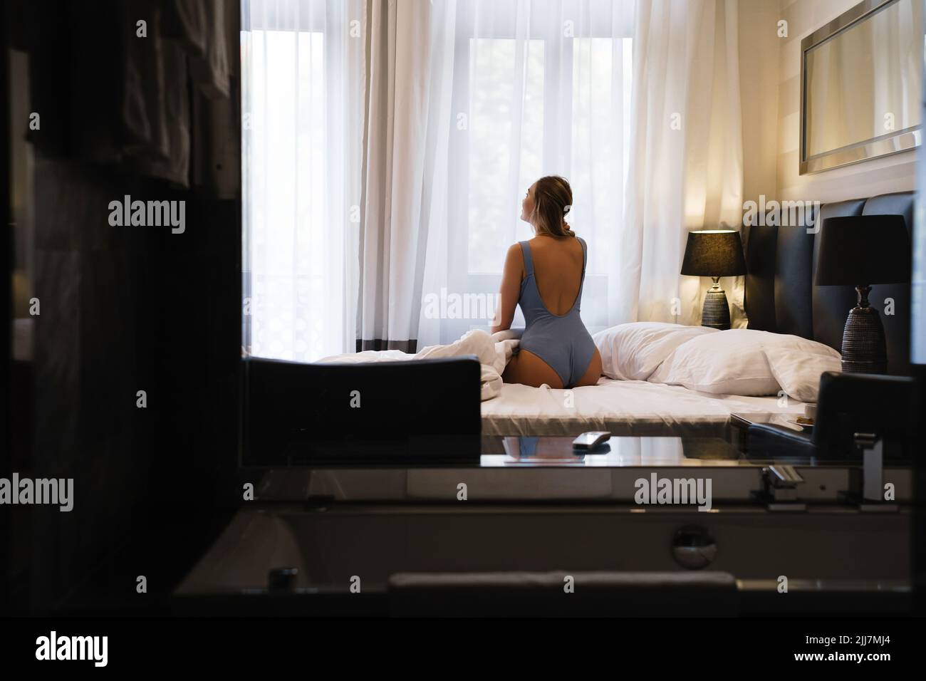 Donna attraente in camera d'albergo seduta nel letto sopra la finestra dal retro, appassionata Donna in tuta avendo riposo in hotel moderno durante il viaggio Foto Stock