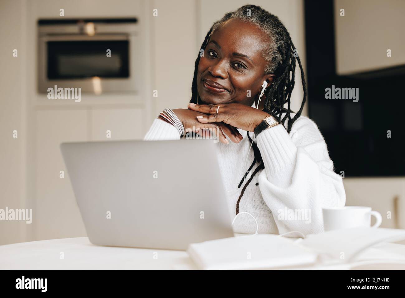 Donna d'affari etnica che guarda la macchina fotografica mentre lavora su un laptop nel suo ufficio domestico. Donna d'affari matura che ascolta musica mentre lavora da ho Foto Stock