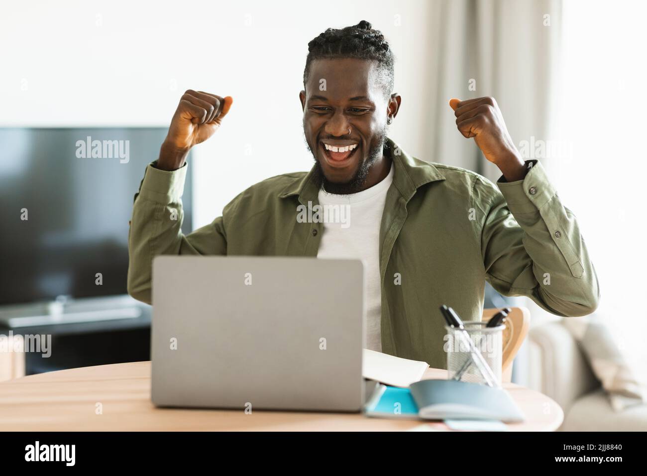 L'uomo nero ipergioioso gesturando sì davanti al portatile, celebrando il successo, seduto al tavolo in ufficio Foto Stock