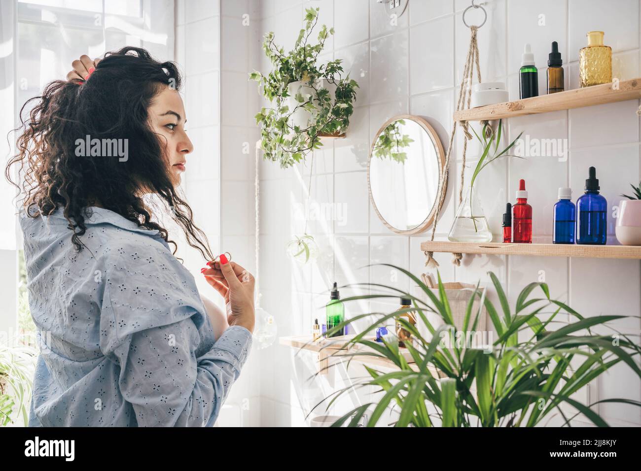 Giovane donna di brunette sovrappeso con capelli ricci che guarda in mirrow in bagno chiaro con shalves di legno per cosmetici naturali. Concetto di benessere e positività del corpo Foto Stock