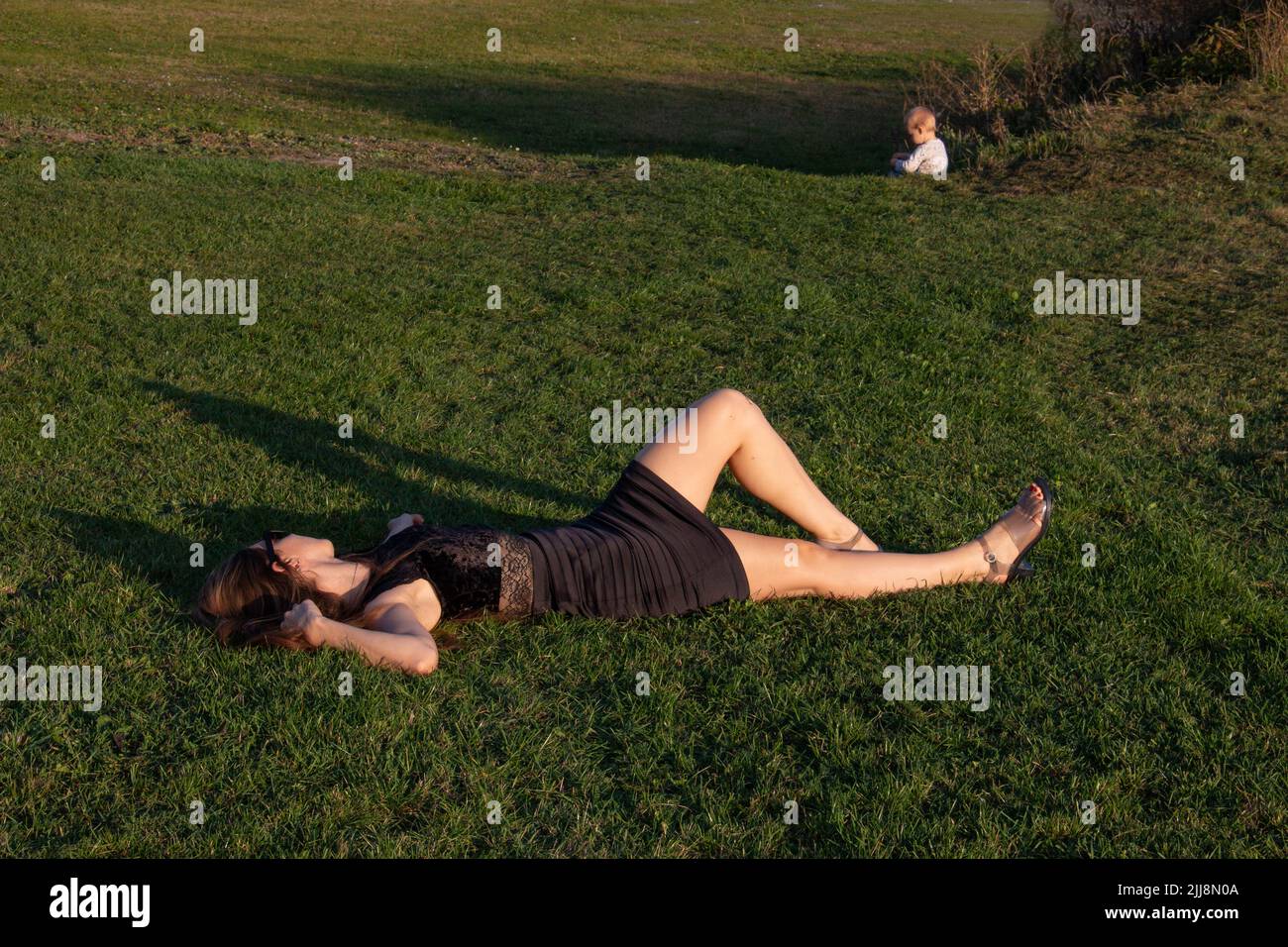 Madre e bambino fuori, madre esausta che riposa mentre il bambino gioca sull'erba Foto Stock