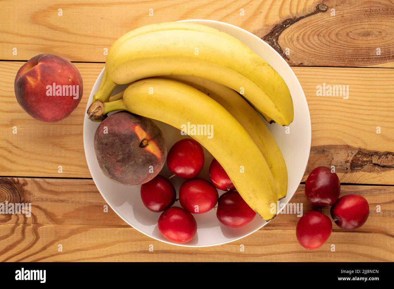 Alcune susine dolci di ciliegia succosa, due pesche e un mazzo di banane con utensili in ceramica bianca su un tavolo di legno, primo piano, vista dall'alto Foto Stock