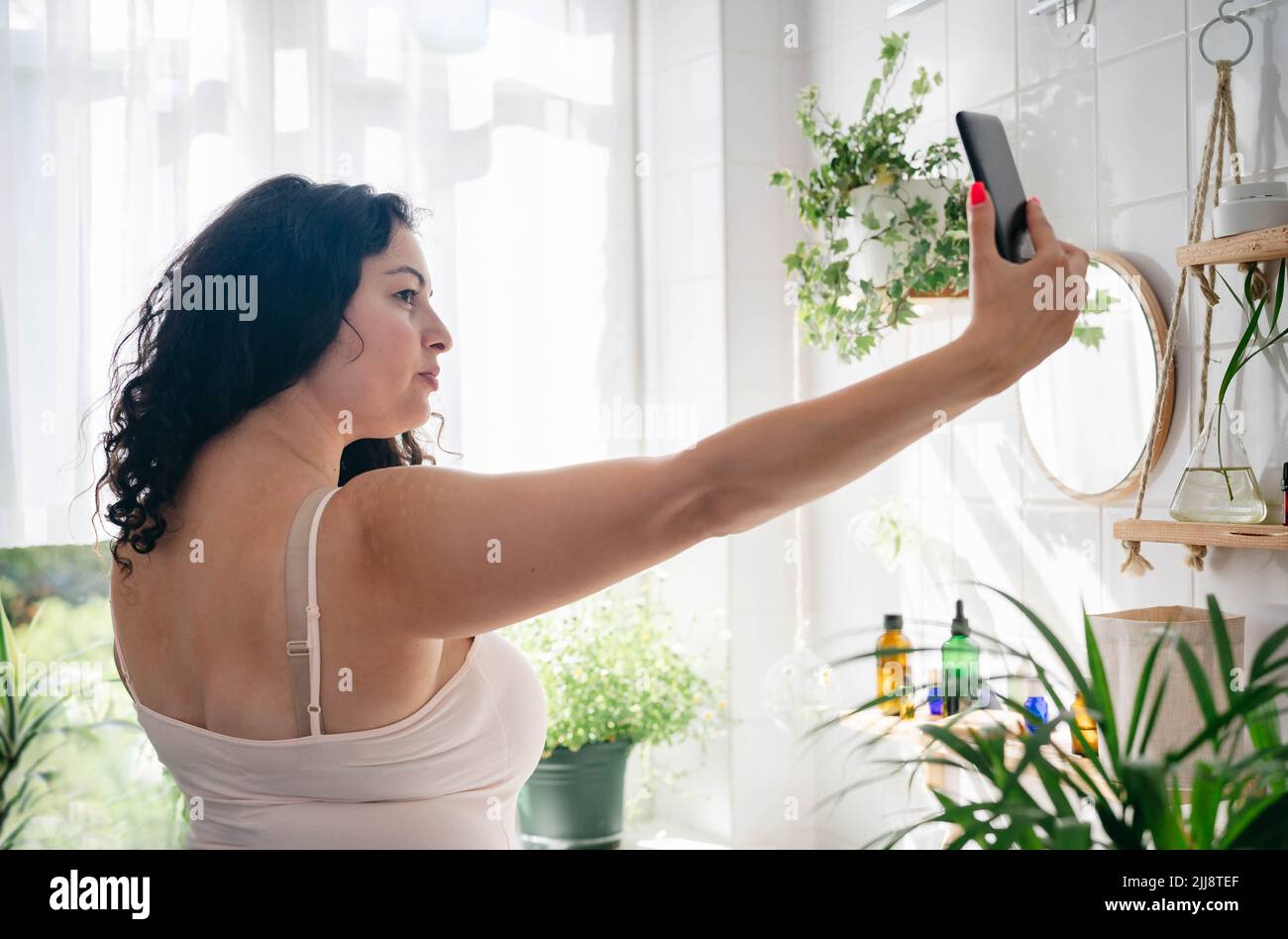 Donna in intimo rosa con selfie in bagno. Benessere e tecnologia quotidiana. Positività del corpo. Zona comfort Foto Stock