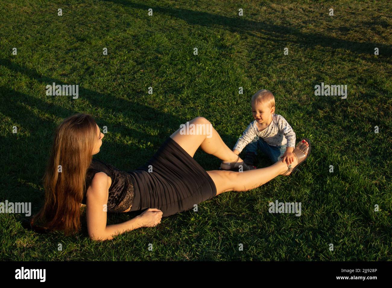 Figlio piccolo del toddler che gioca con le gambe della madre, donna con il suo bambino all'aperto sul prato dell'erba Foto Stock