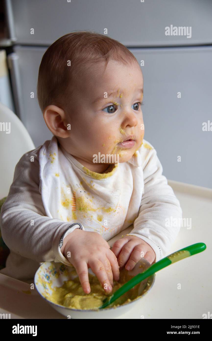 Bambino sudicio e sporco che mangia cibo con le mani sul seggiolone, ragazzo che impara a mangiare Foto Stock