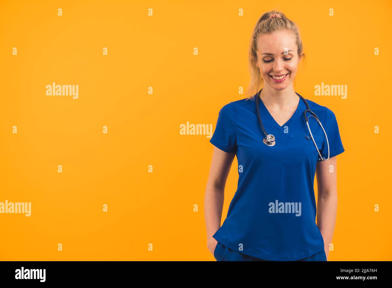 Bella bionda sottile donna caucasica in copia spazio su sfondo arancione indossando blu scuro uniforme ospedale e stetoscopio. Espressione facciale sorridente, guardando verso il basso. Foto di alta qualità Foto Stock
