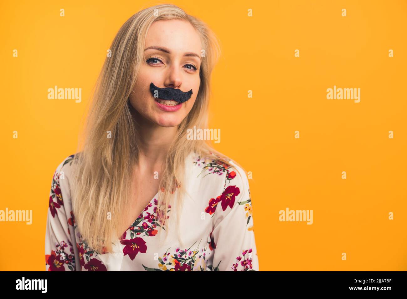 Concetto di Movember. Spazio copia, foto studio, isolato su sfondo arancione. Funky bella ragazza caucasica con capelli biondi in una blusa floreale bianca in posa per fotocamera con falsi baffi neri sotto il naso. Foto di alta qualità Foto Stock