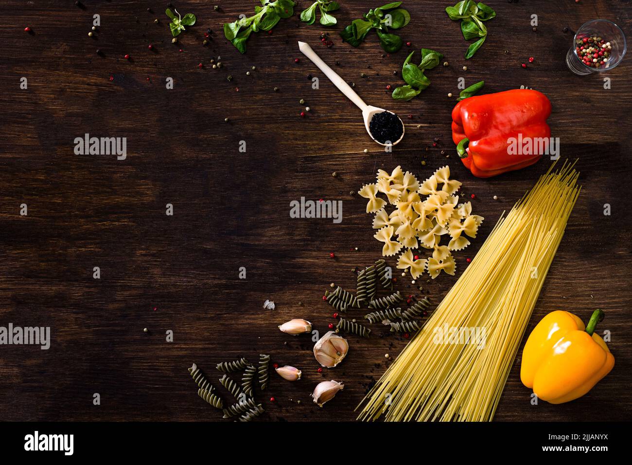 Vari tipi di pasta su sfondo di legno scuro. FARFALLE, fusilli, spaghetti, peperone, insalata di mais, spicchi d'aglio rossi, verdi o gialli Foto Stock