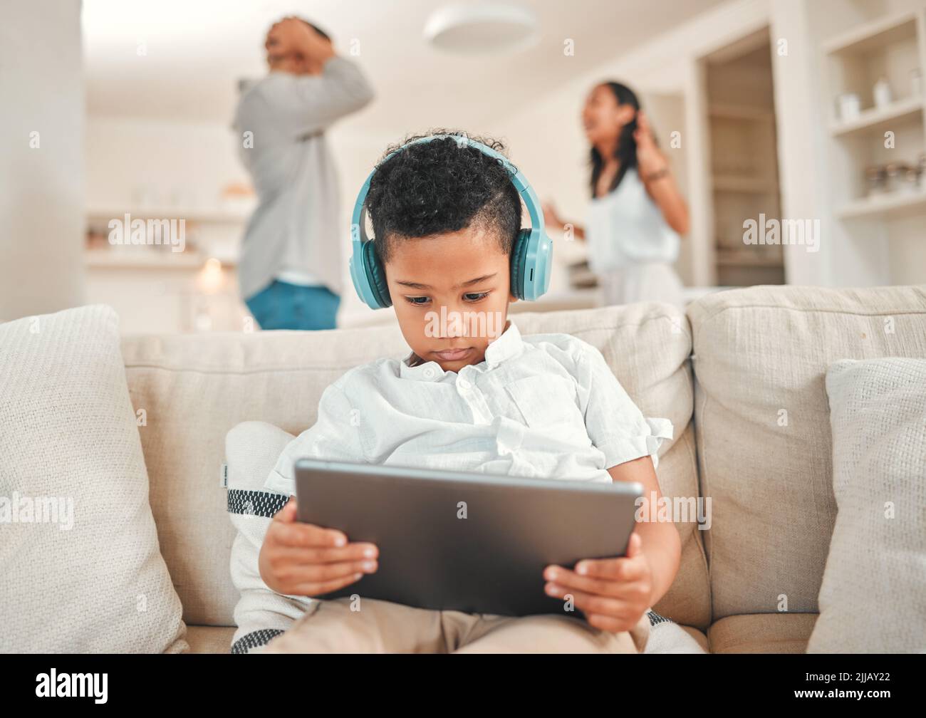 Non riesco più a vivere in questo modo. Un ragazzino che indossa una cuffia e usa un tablet digitale mentre i suoi genitori discutono in background a casa. Foto Stock