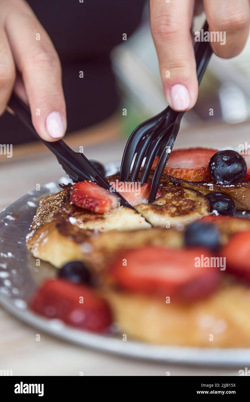 Mani che usano argenteria di plastica per tagliare i pancake di mirtillo e fragola in un ristorante Foto Stock
