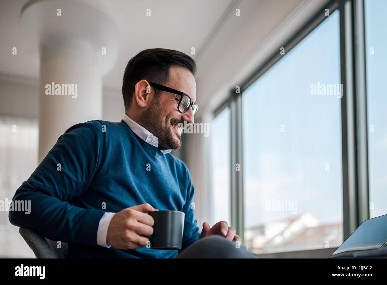 Sorridente uomo d'affari che ha un caffè mentre lavora. Il giovane professionista di sesso maschile è seduto alla scrivania. Indossa astuti casuals in ufficio. Foto Stock
