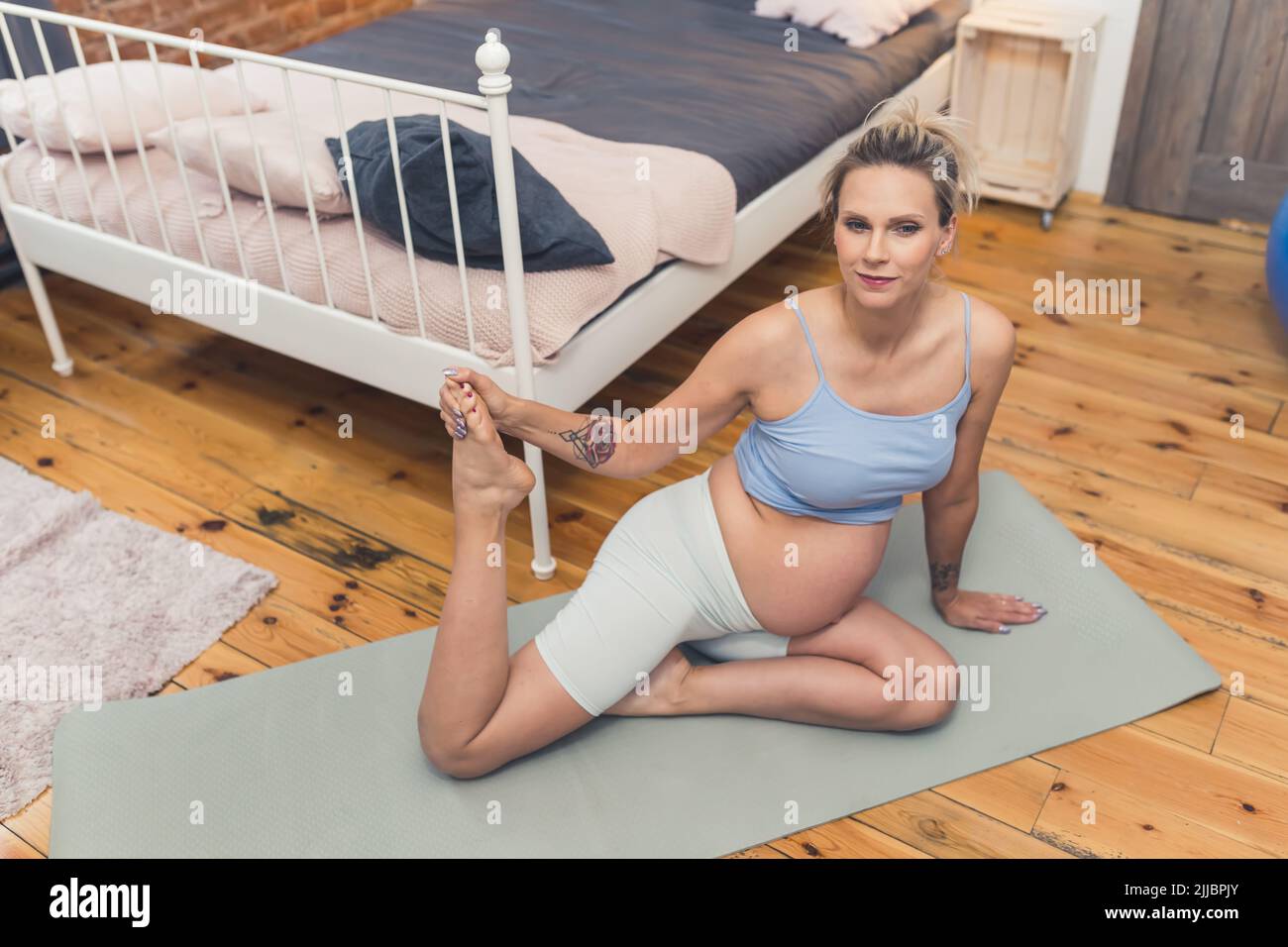 Stretching durante la gravidanza concetto. Donna incinta europea bionda di mezza età che allunga la coscia mentre si siede su un tappeto yoga. Scatto al coperto. Foto di alta qualità Foto Stock