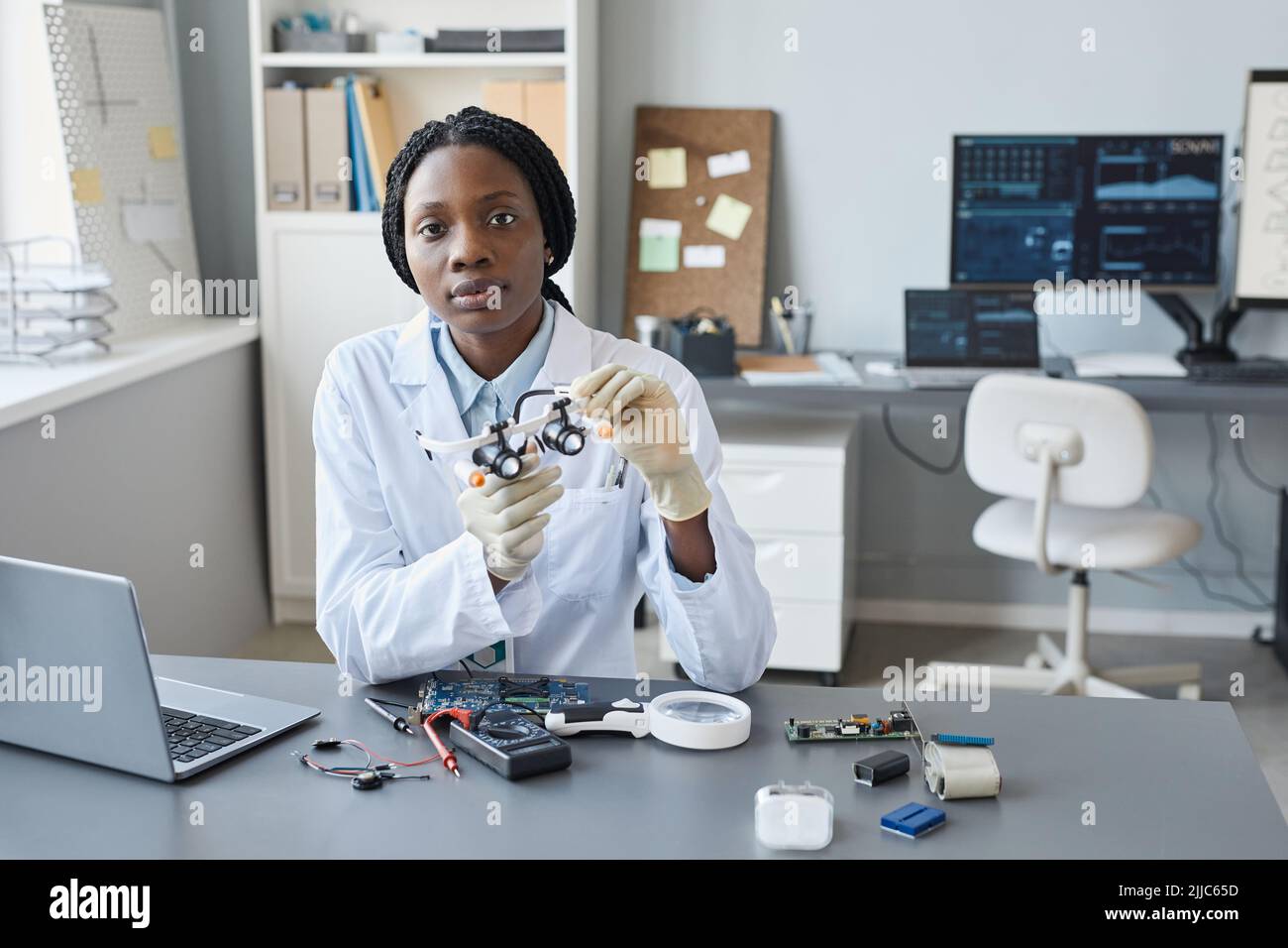 Ritratto di giovane scienziata femminile che guarda la fotocamera mentre si siede sul posto di lavoro in laboratorio di ingegneria, spazio copia Foto Stock