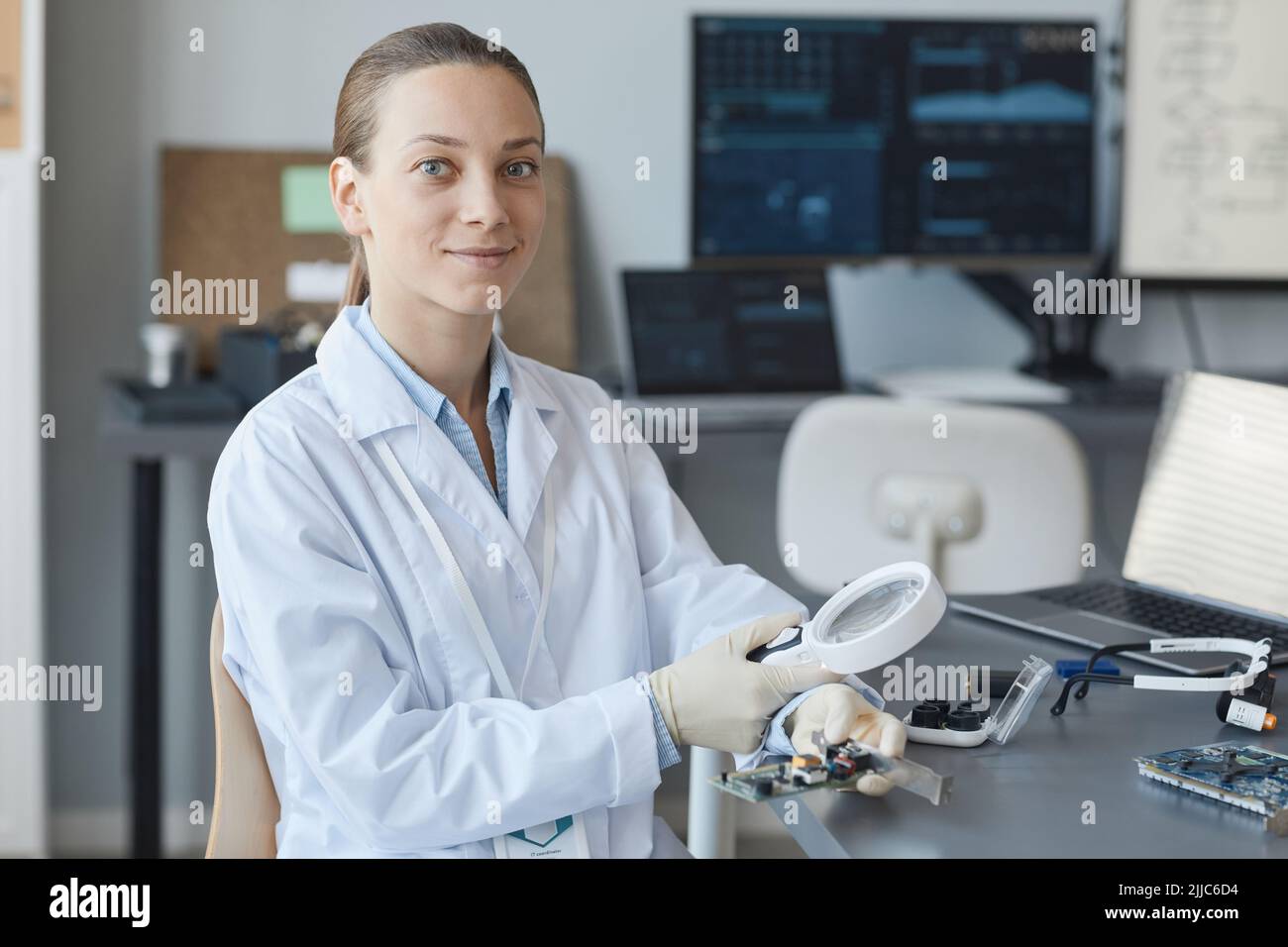 Ritratto di giovane scienziata femminile sorridente alla macchina fotografica mentre si siede sul posto di lavoro in laboratorio di ingegneria, spazio copia Foto Stock