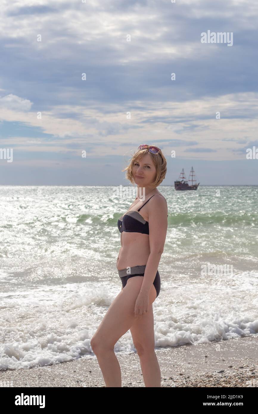 Donna snella matura in costume da bagno sullo sfondo del mare con una nave. Viaggi e turismo, vacanze e relax. Foto Stock