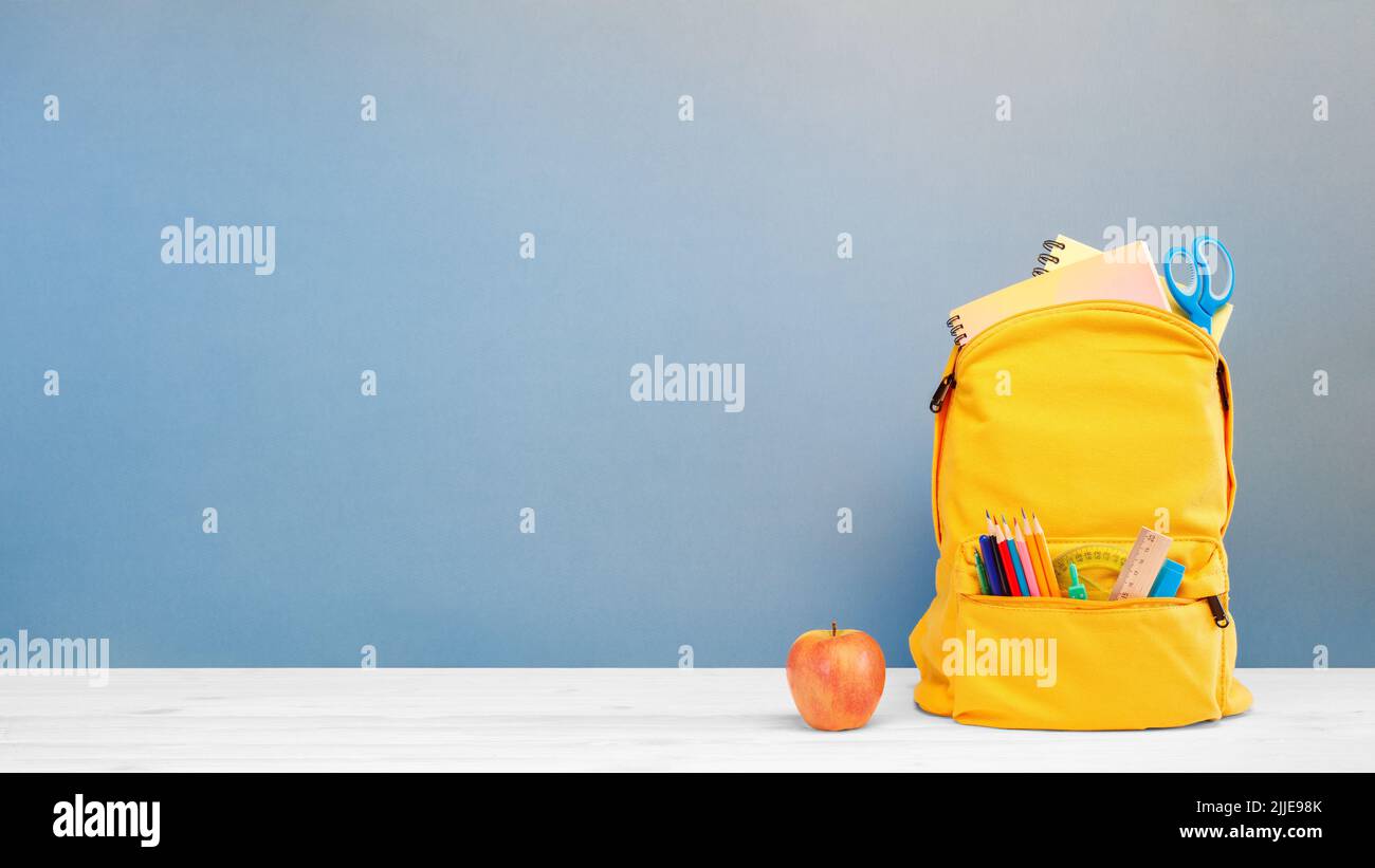 Zaino con diverse cancelleria colorata e mela sul tavolo e sfondo blu. Banner design back to School concept background. Foto Stock