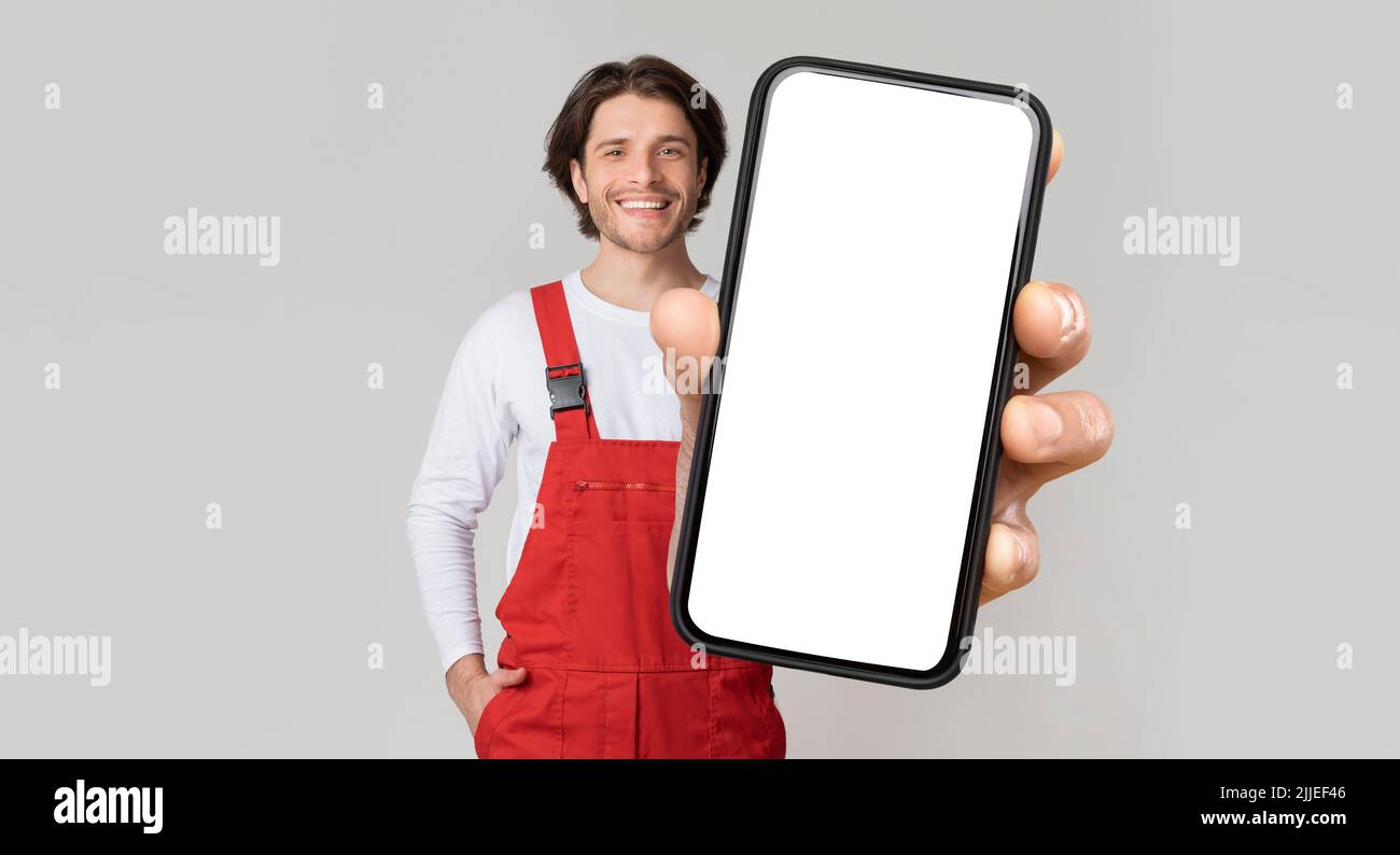 Concetto di Handyman Services. Un giovane uomo di lavoro sorridente in uniforme che mostra lo smartphone Foto Stock