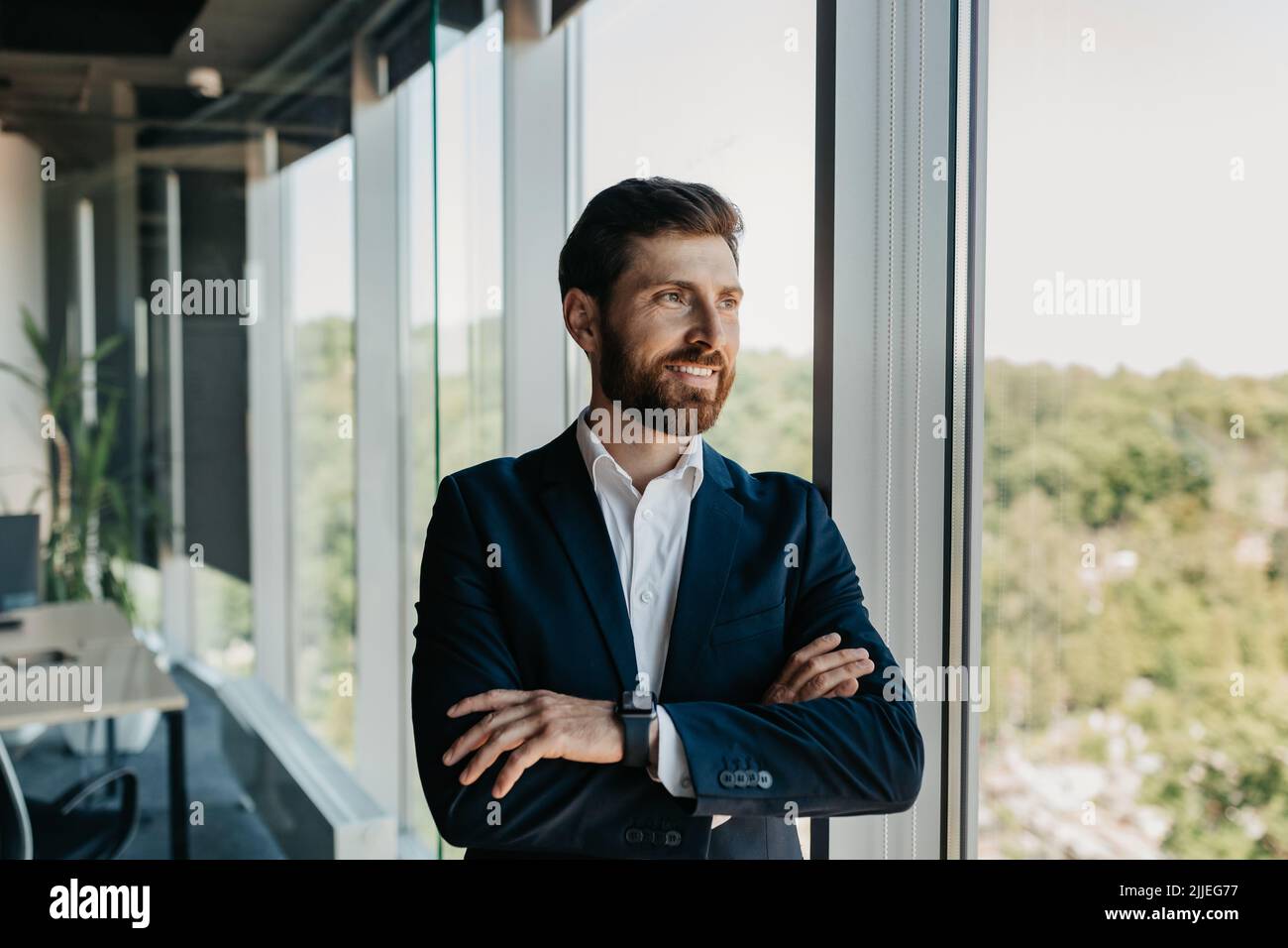 Ritratto di bell'uomo d'affari caucasico in piedi con le braccia ripiegate vicino alla finestra, dopo una pausa in ufficio, spazio libero Foto Stock