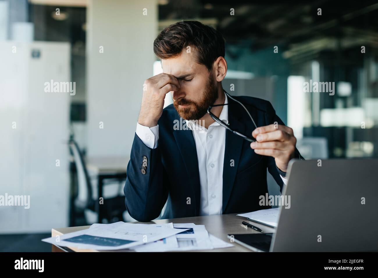 Uomo d'affari esausto seduto davanti al computer portatile in ufficio moderno, tenendo gli occhiali e strofinando ponte naso Foto Stock
