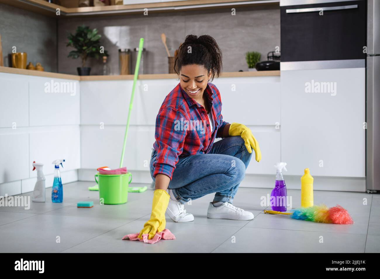 Felice millenaria donna afroamericana casalinga in guanti di gomma lavare pavimento con forniture di pulizia Foto Stock