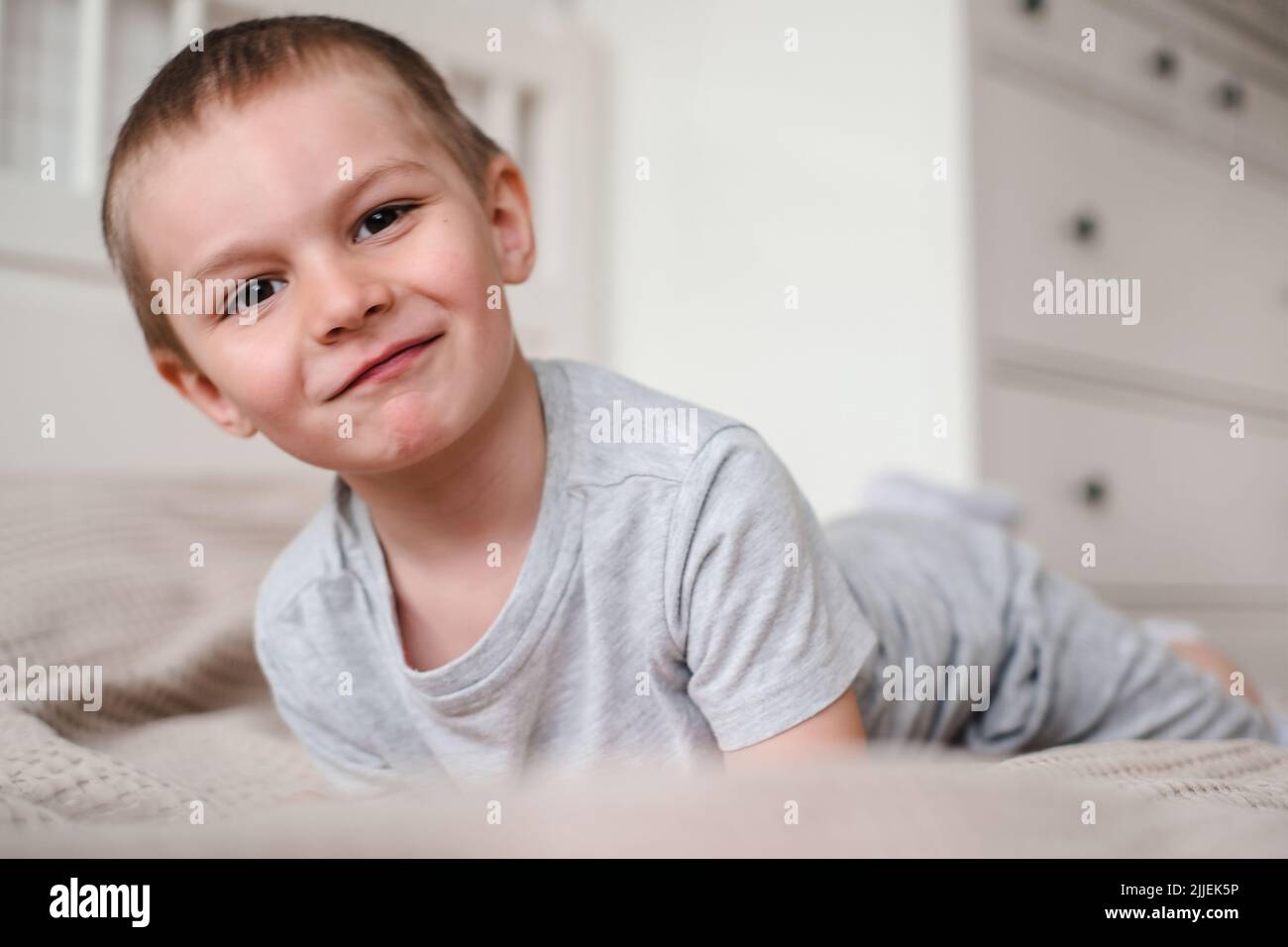 Primo piano di un piccolo ragazzo caucasico felice in una t-shirt grigia si trova sul letto e sorride Foto Stock